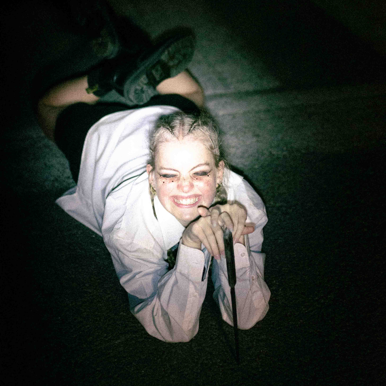 Projet Court Métrange : photo d'une fille aux yeux maquillés de noir, avec des tresses blondes, allongée par terre qui sourit de manière insolente et qui tient un couteau à la main